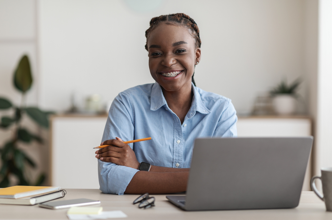 Woman in a blue collared shirt sitting at a desk behind a laptop smiling.